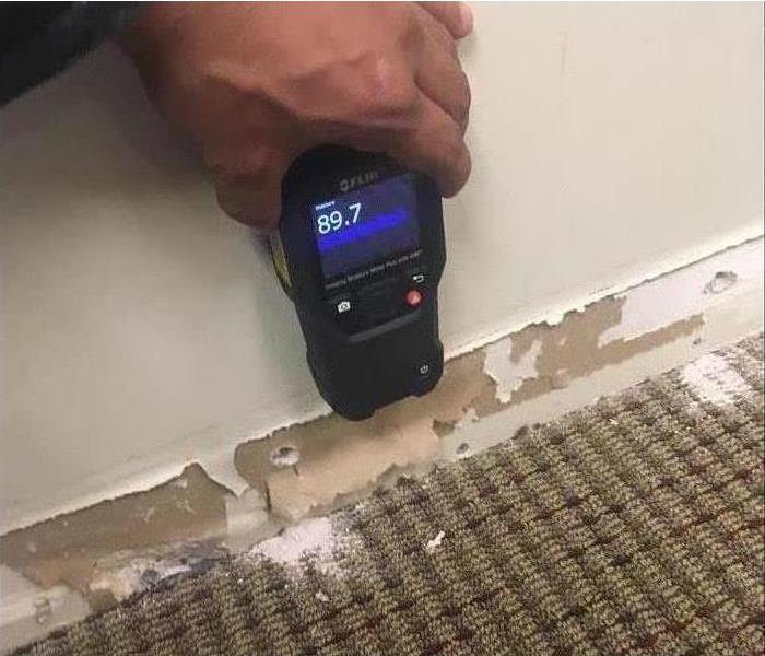 A hand holding a moisture meters against a wall and baseboards are removed due to water damage 